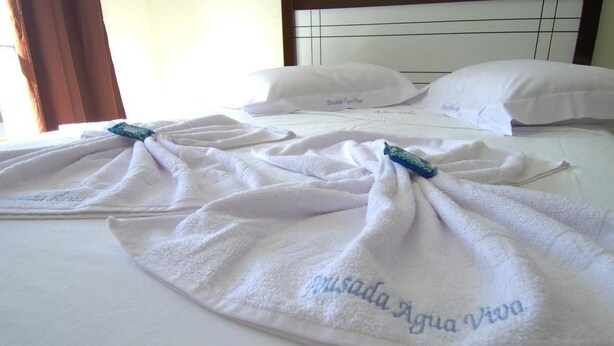 Una habitación de hotel con una cama hecha con esmero, toallas blancas con texto bordado y una ventana con cortinas.