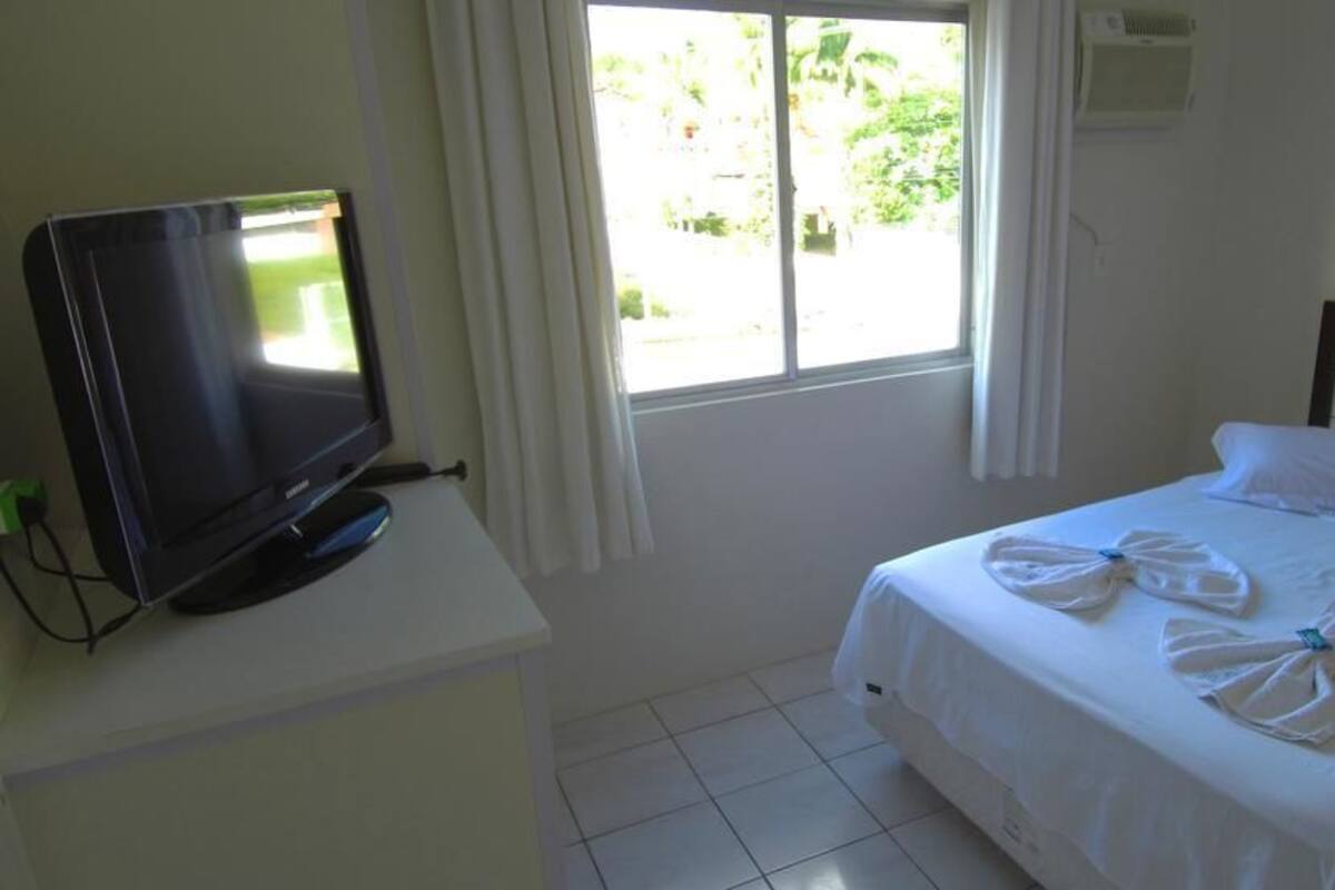 Quarto de hotel com uma cama, uma televisão sobre uma mesa branca e uma janela com cortinas.