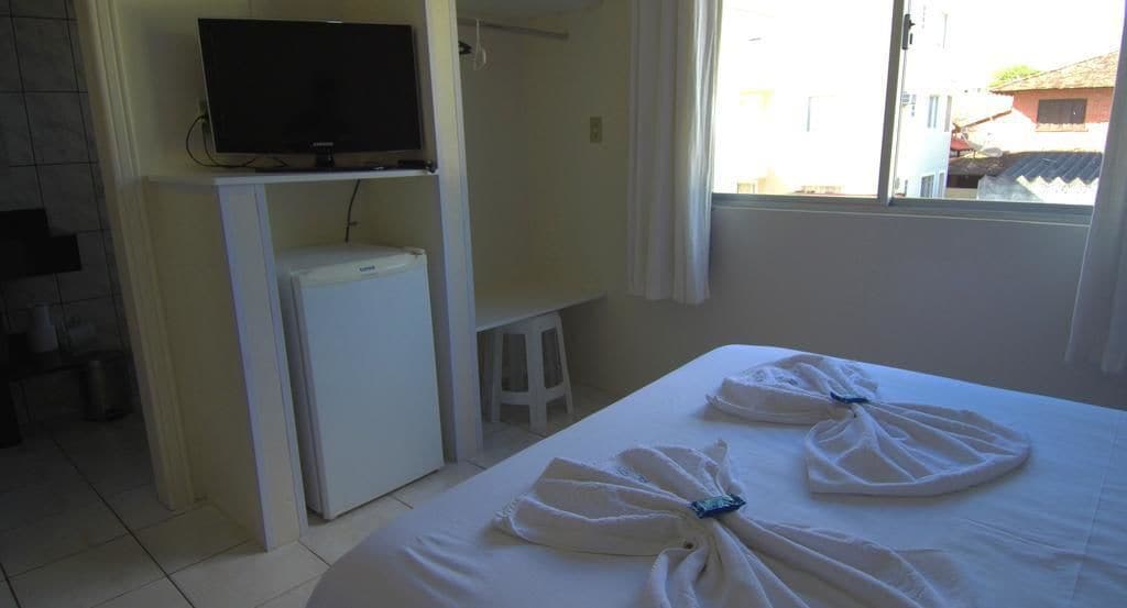 A hotel room with a bed, a television, a refrigerator, and a window with a view of buildings.