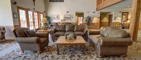 A spacious living room with a high ceiling, wooden ceiling beams, and a large window. There are three leather sofas, a wooden coffee table, and a patterned rug.