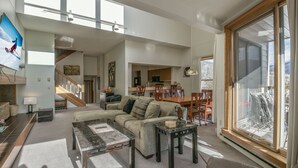 En rummelig opholdsstue med en stor sofa, et sofabord og et spisebord. Der er en trappe til en etage ovenpå, og et stort vindue giver naturligt lys.