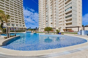 A swimming pool surrounded by high-rise buildings with balconies, a clear sky, and some palm trees. - APOLO VXI ONE BEDROOM APARTMENT WITH SEAVIEWS (Calpe / Calp)