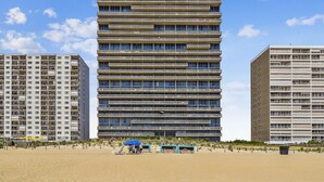 Un edificio de varios pisos con fachada plana, situado junto a una playa de arena con algunas sillas y sombrillas.