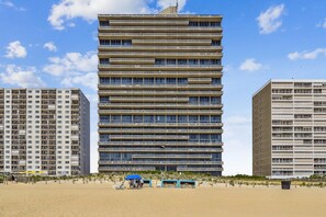 Un edificio a più piani con facciata piatta, situato accanto a una spiaggia sabbiosa con alcuni lettini e ombrelloni.