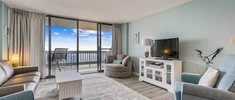 Una sala de estar con sofá, sillón, televisor y vista al océano a través de un ventanal corredero.