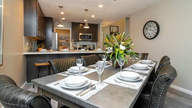 A dining area with a long table set for four, surrounded by leather chairs. There is a kitchen in the background with modern appliances and a clock on the wall. - At Base of Cottonwoods! * Private Hot Tub * Xbox (Cottonwood Heights)