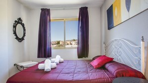 A bedroom with a bed, a window with curtains, a bedside table, and a view of buildings outside. - MARE NOSTRUM - Apartment near the beach with private pool (Calp)
