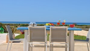 Una mesa con sillas orientada a una vista panorámica del mar, un bol de fruta y dos copas de vino tinto.