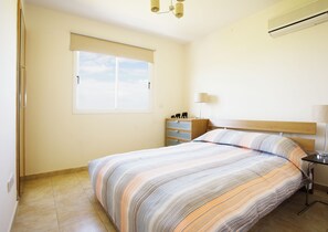 Une chambre à coucher comprenant un lit, une table de chevet, une lampe, une commode et une fenêtre avec des stores.