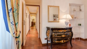 Un pasillo con cortinas estampadas, suelo de madera, un aparador negro decorado con motivos florales, un espejo y un estante blanco con objetos.
