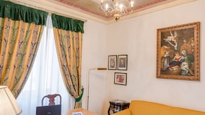 Una habitación con suelo de madera, un sofá amarillo, un escritorio con lámpara, una araña y un cuadro en la pared.