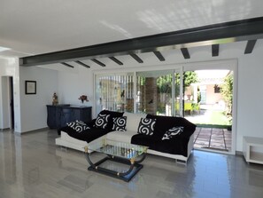 A modern living room with a glass coffee table, a white sofa with black patterned cushions, and a view of the outdoor garden through a sliding glass door.