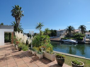 Um imóvel à beira-mar com um pátio pavimentado, plantas em vasos, um barco atracado e arquitetura tradicional ao fundo.
