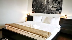 A neatly made bed with white linens and a brown throw blanket, flanked by two bedside tables with lamps, in a room with a cityscape view. - Studio G by MRG Apartments | Steps away from Metro & Amenities (București)