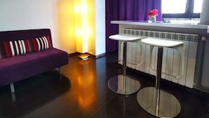 A modern living room with a purple sofa, two white bar stools, a floor lamp, and a window with purple curtains. - Studio G by MRG Apartments | Steps away from Metro & Amenities (București)