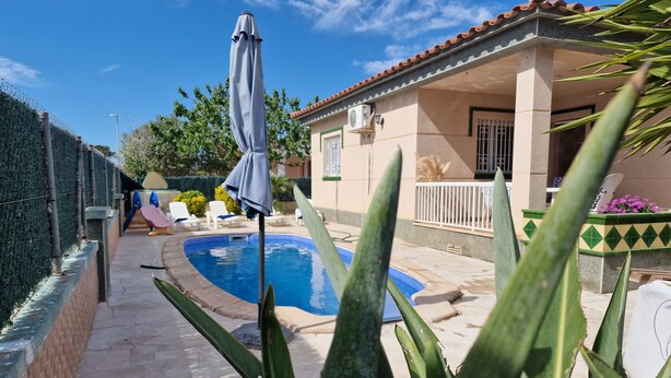 Uma área de piscina com espaço de estar coberto, jardim cercado e um edifício com telhado em azulejo.