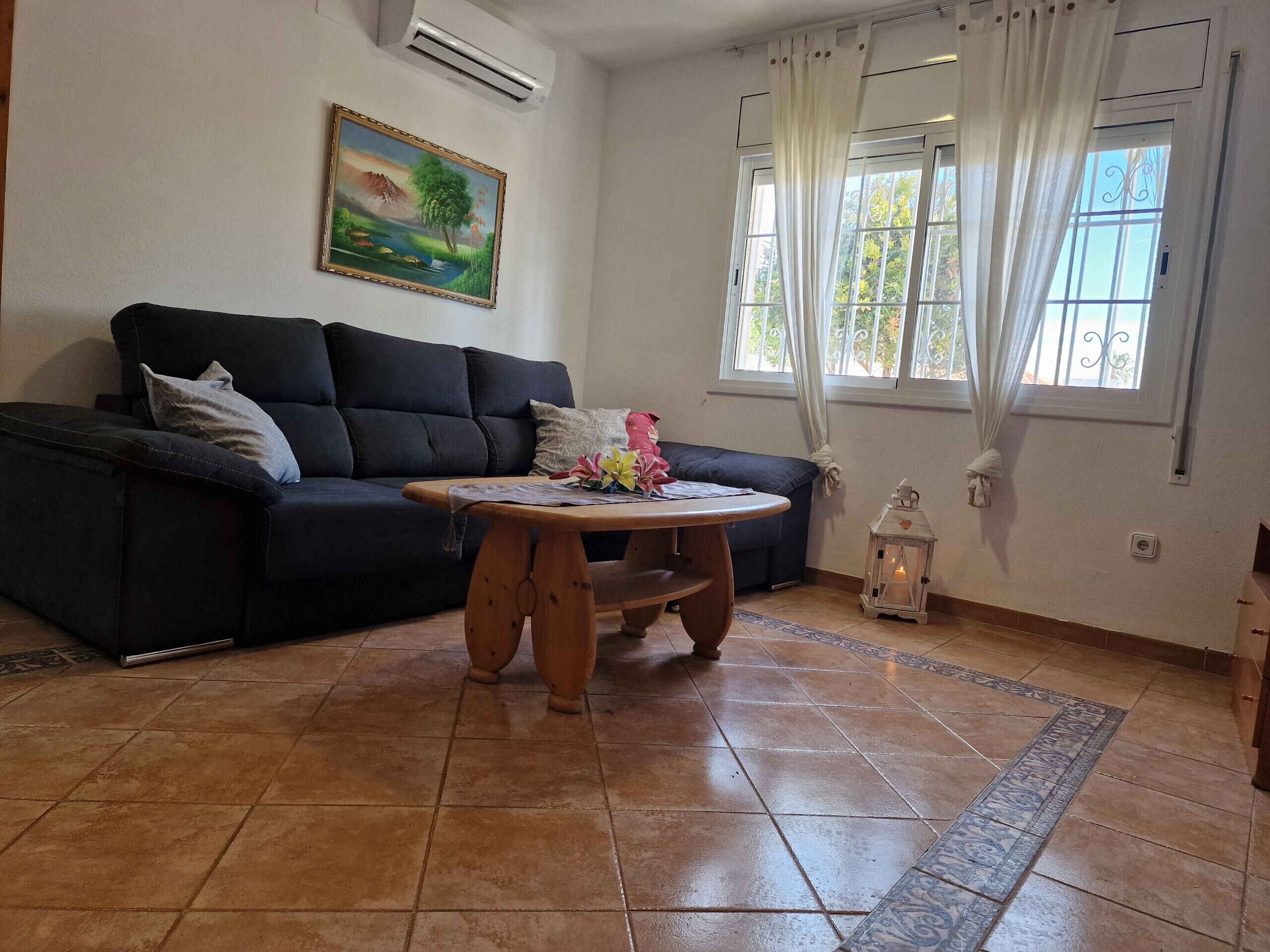 Una sala de estar con un sofá oscuro, una mesa de centro de madera, un cuadro en la pared, una ventana con cortinas y un suelo de baldosas.