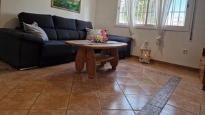 Uma sala de estar com um sofá escuro, uma mesa de centro de madeira, um quadro na parede, uma janela com cortinas e um chão de azulejos.