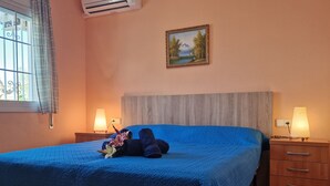 Quarto com colcha azul, cabeceira de madeira, duas mesas de cabeceira com abajures, um quadro na parede e uma janela com cortinas.