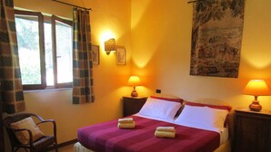 Camera da letto con un letto, due lampade ai comodini, una sedia, una finestra con tende e un quadro appeso al muro.