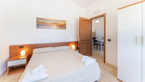 A neatly made bed with towels, a bedside lamp, and a painting on the wall. - One bedroom apartment premium (4 people) - Residence Le Fontane (Villasimius)