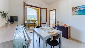 A dining area with a table set for two, a TV on the left, and a view of the outdoors through an open door. - One bedroom apartment premium (4 people) - Residence Le Fontane (Villasimius)