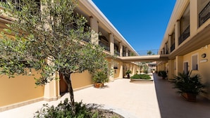 A courtyard with a central tree, potted plants, and a covered walkway connecting multiple buildings. - One bedroom apartment premium (4 people) - Residence Le Fontane (Villasimius)
