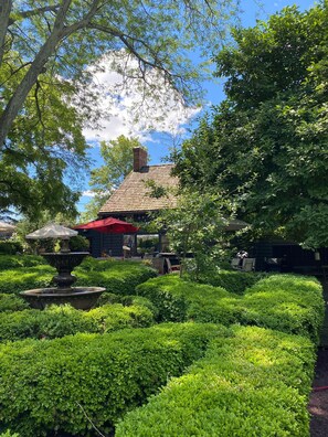 Un jardin agrémenté d’une fontaine, de buissons taillés avec soin et d’un bâtiment surmonté d’un toit rouge.