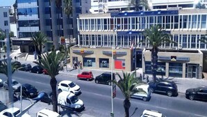 Vista de la calle con un edificio de varios pisos, palmeras y coches aparcados.