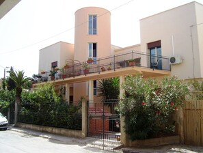 Ein zweistöckiges Gebäude mit Balkon, umgeben von Grünpflanzen und einem verschlossenen Eingang.