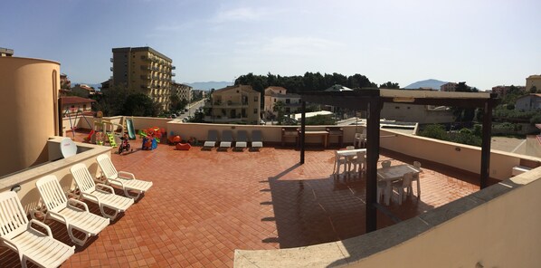 Eine Dachterrasse mit weißen Liegestühlen, einer überdachten Lounge und Blick auf umliegende Gebäude und Berge.