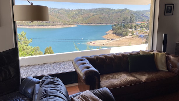 A room with a large window offering a view of a lake and surrounding hills. There is a leather sofa and a lamp near the window. - House in Castelo de Bode, blue lake (Castelo de Bode)