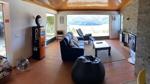 A spacious living area with a wooden ceiling, a large window offering a view of the mountains, a sofa, a bean bag, and a television. - House in Castelo de Bode, blue lake (Castelo de Bode)
