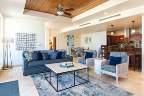 A modern living room with a sofa, coffee table, and wicker chairs, a dining area with chairs, and a kitchen in the background.