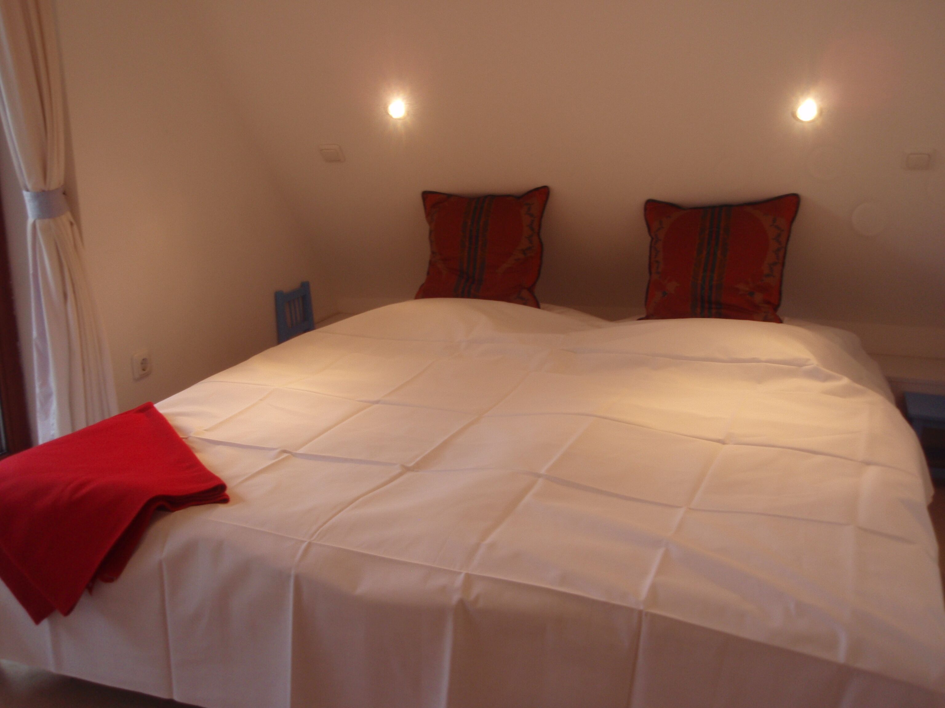 Una cama bien tendida con edredón blanco y almohadas rojas, una manta roja y una silla azul en una habitación sencilla con paredes y luces de techo blancas.