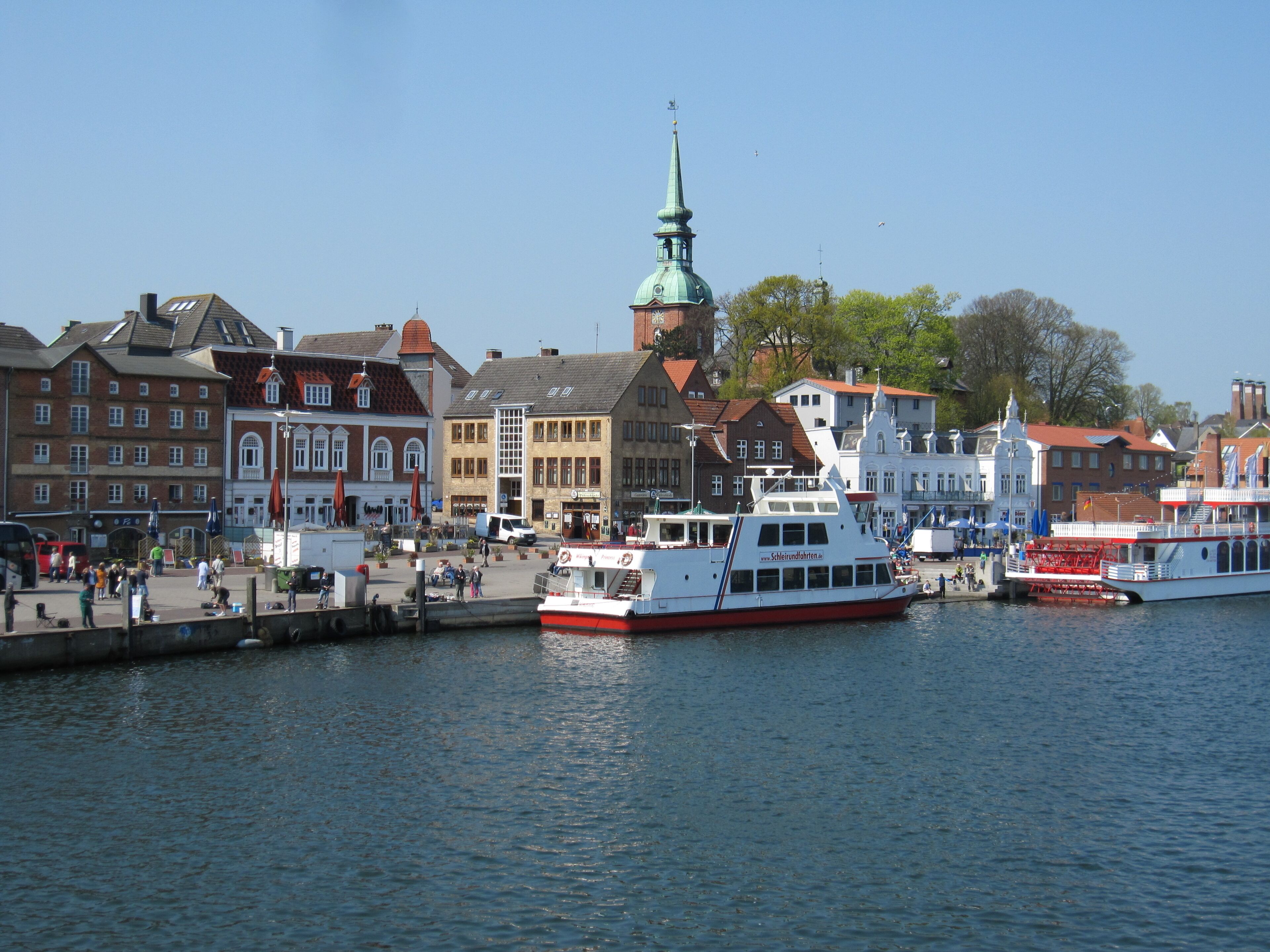 A waterfront area with historic buildings, a prominent church tower, and a large white boat docked at the pier.