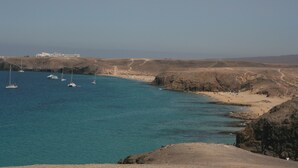 Un paysage côtier avec une eau d’un bleu limpide, un terrain rocheux et un bâtiment visible au loin.