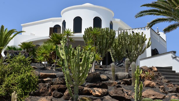 Un bâtiment blanc doté de fenêtres en arc de cercle et d’un toit en forme de dôme, entouré de cactus et de rochers.