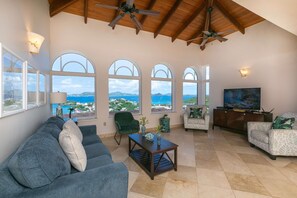 Un salon spacieux avec un grand canapé, des fauteuils, une table basse et un téléviseur. De grandes fenêtres en arc offrent une vue sur la mer et les montagnes.
