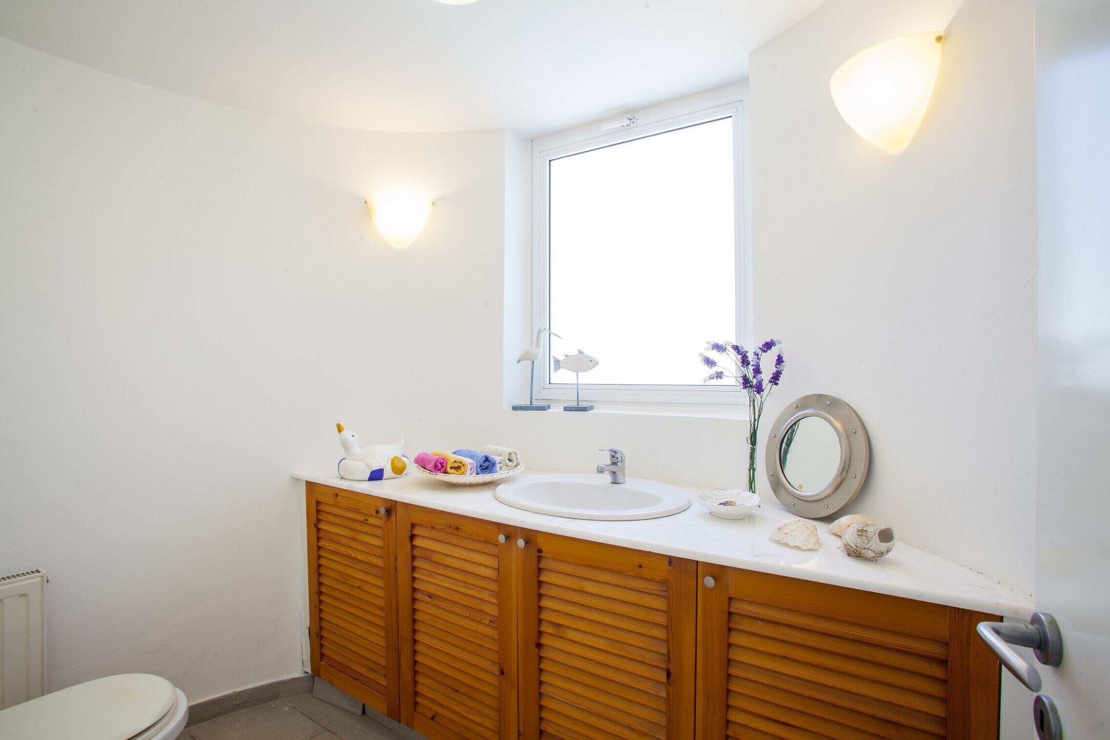 Un baño con lavamanos blanco, armarios de madera, un espejo y una ventana.