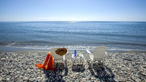 Une plage avec deux chaises blanches et une petite table, un chapeau de paille et un tissu orange sur le rivage de galets.