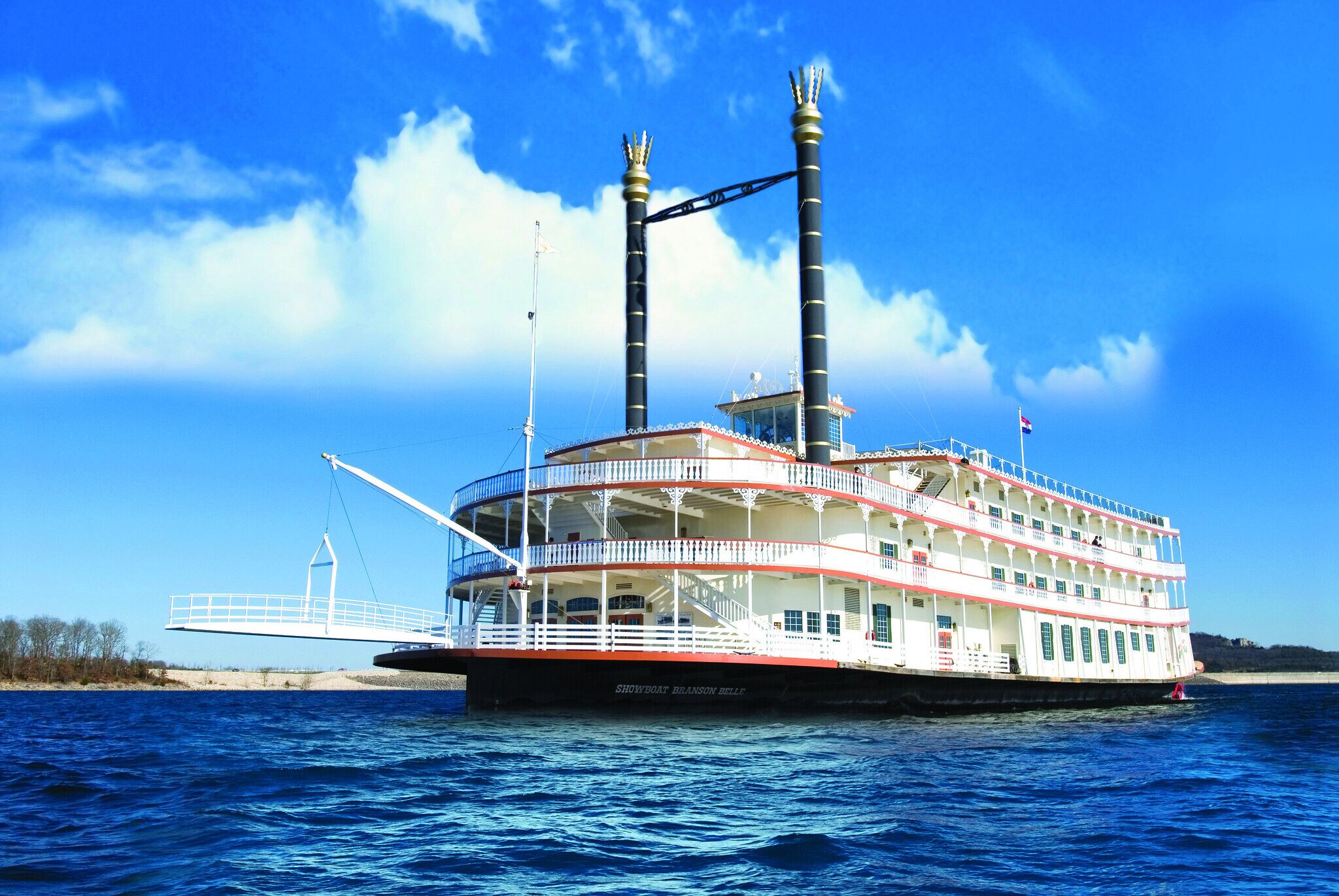 A large, multi-deck riverboat with a prominent chimney structure, a white and red exterior, and a flag on top.