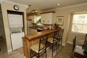 A kitchen with a wooden bar, white cabinets, a clock, and a window with blinds.