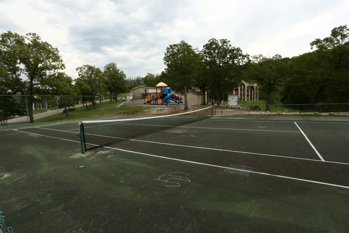 Un court de tennis en plein air, doté d’un filet, entouré d’arbres et avec une aire de jeux en arrière-plan.