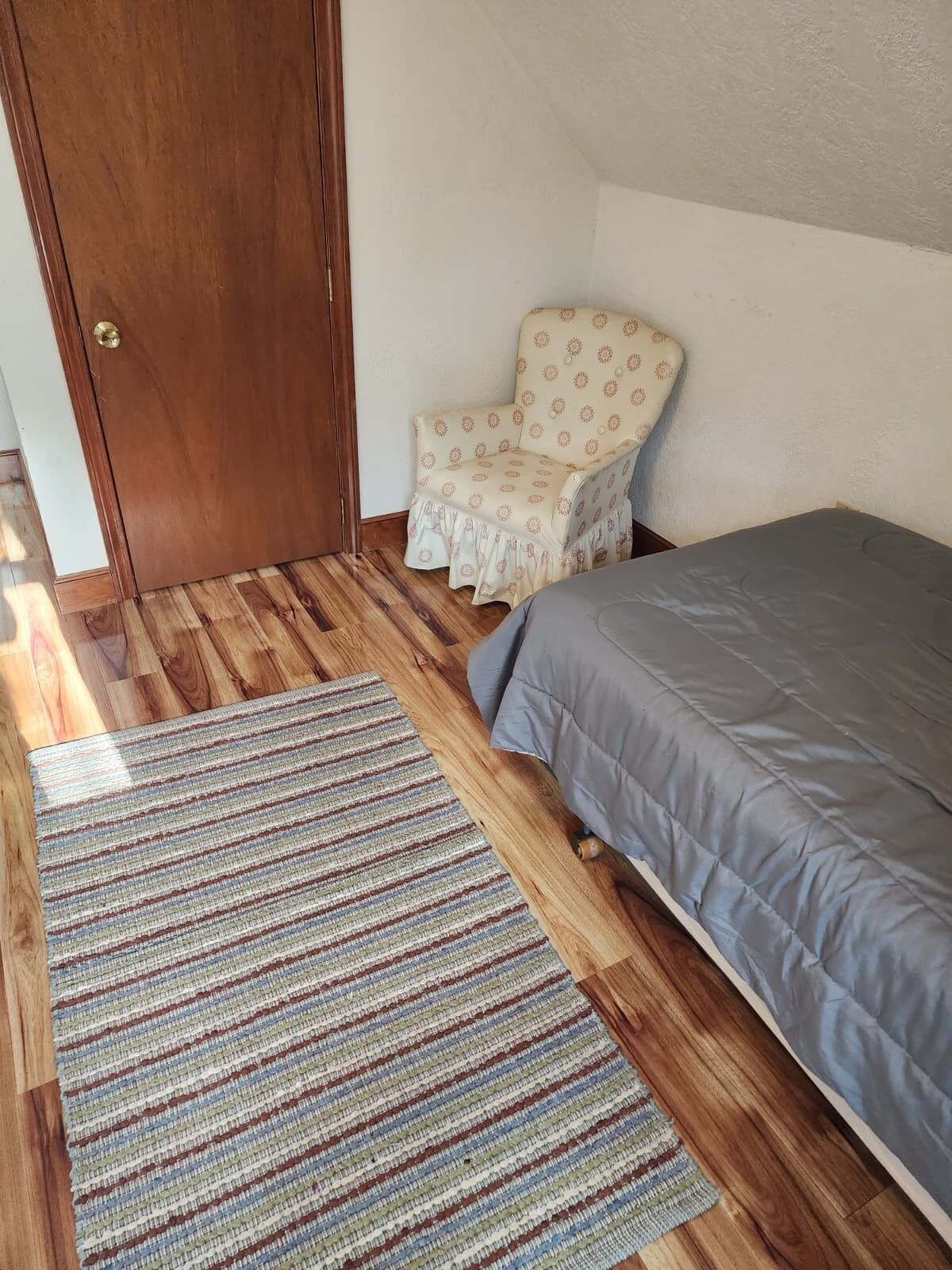 Una habitación con piso de madera, una cama con cubrecama gris, un tapete a rayas y un sillón con estampado.