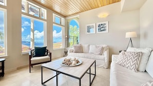 A spacious living room with a wooden ceiling, large windows offering a view of the sea, a white sofa, a coffee table, and a floor lamp. - Frangipani Private Pool and Tesla Backup! (St. John)