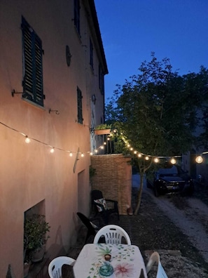 En udendørs terrasse med bord og stole, lyskæder og en parkeret bil.