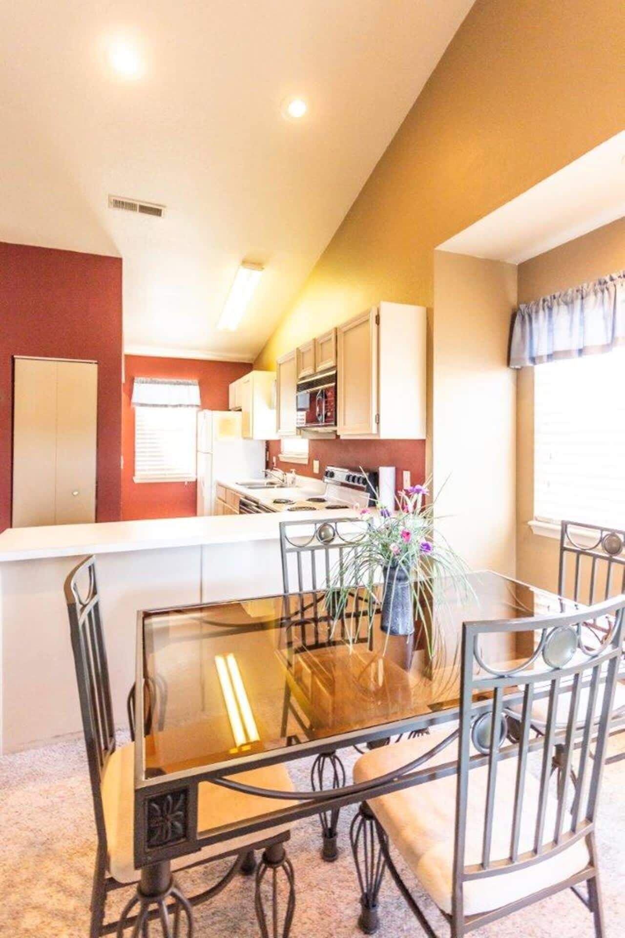 Comedor con mesa de vidrio y sillas de metal, cocina al fondo y ventana con cortinas.