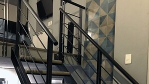 Una escalera con barandillas negras y peldaños de madera, que conduce a una zona de descanso con un sofá azul y una cocina al fondo.