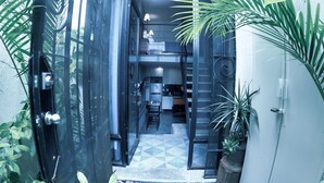 Una entrada moderna y cerrada con puerta de vidrio, un pequeño espacio de cocina visible en el interior, y plantas en macetas alrededor.